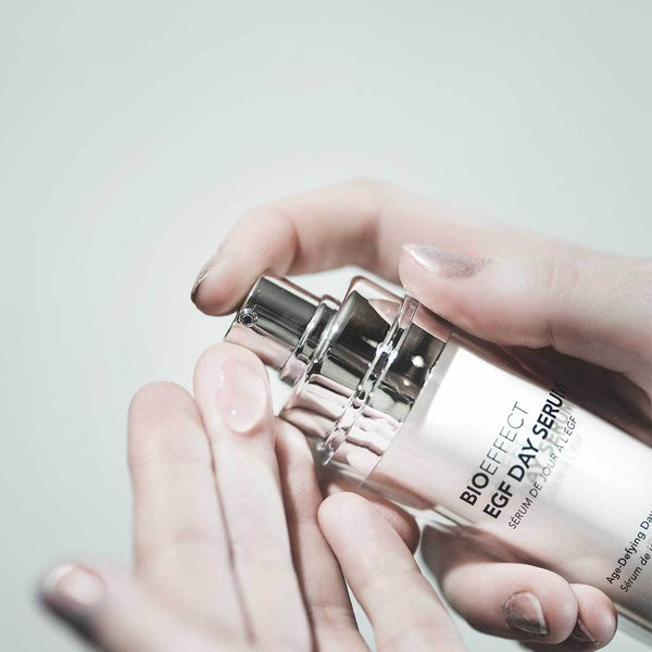 Woman’s hands holding a bottle of BIOEFFECT EGF Day Age-Defying Serum with a silver lid and squeezing a small amount onto her left hand. Her nails are painted with a light silver glitter.
